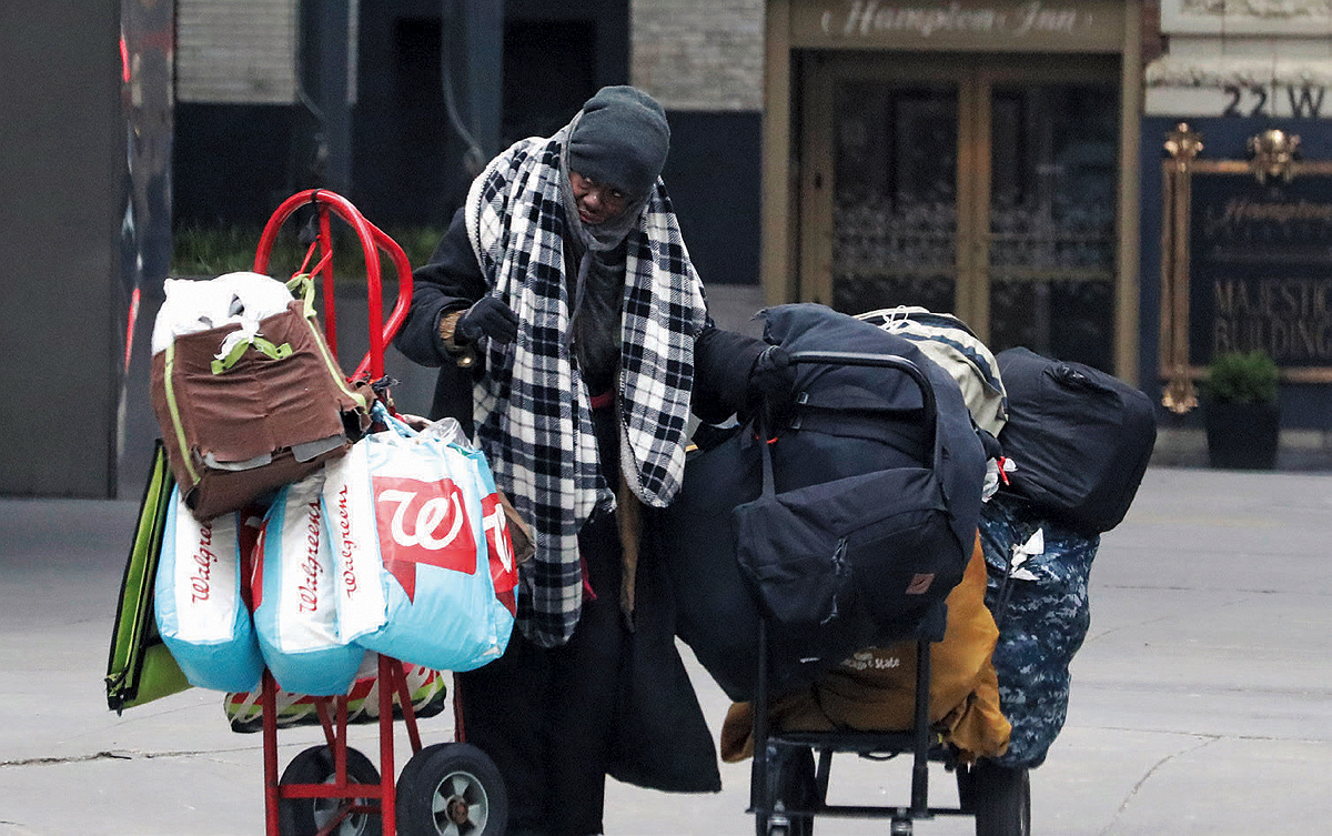 A woman stands on the street in a winter scarf and hat pulls two wheeled dollies loaded with bags of her possessions 