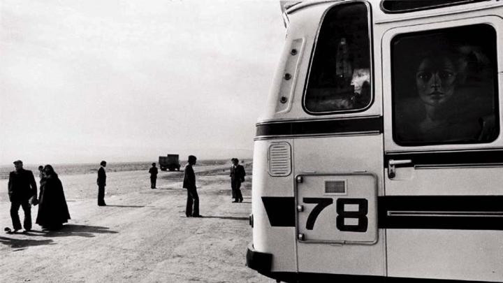 Black-and-white photo of a bus, dominating the right half of the photograph, while a handful of people, and a truck driving toward the horizon, appear as small figures in the left half.